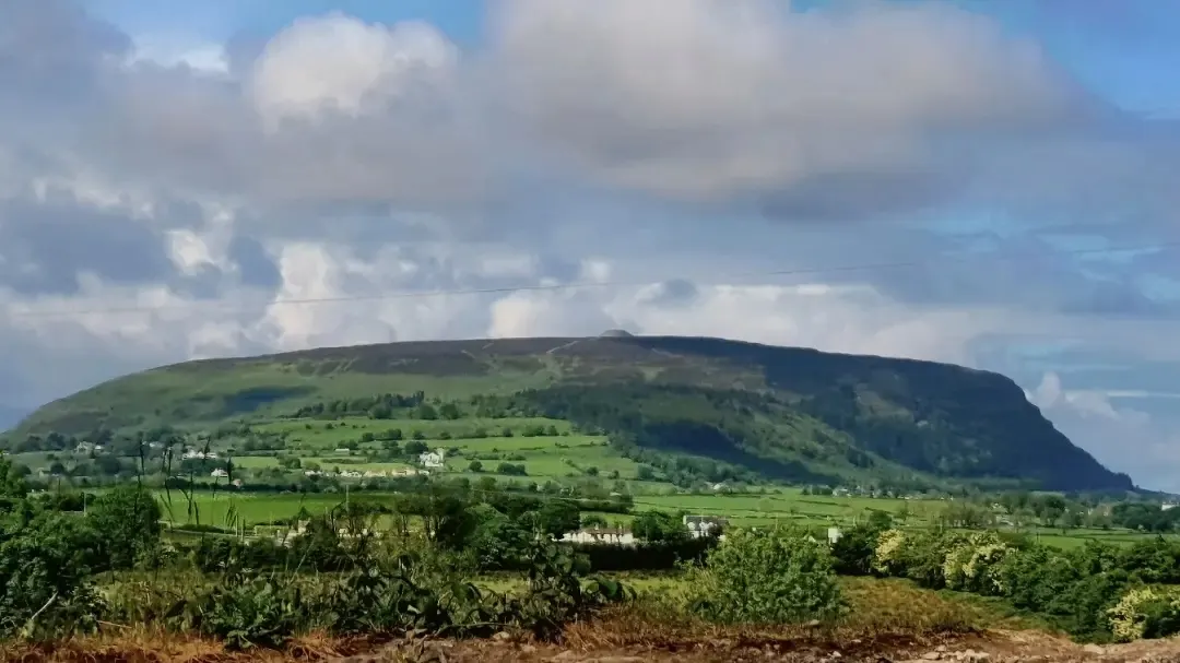 A broad green mountain ridge above fields beneath a cloudy sky.